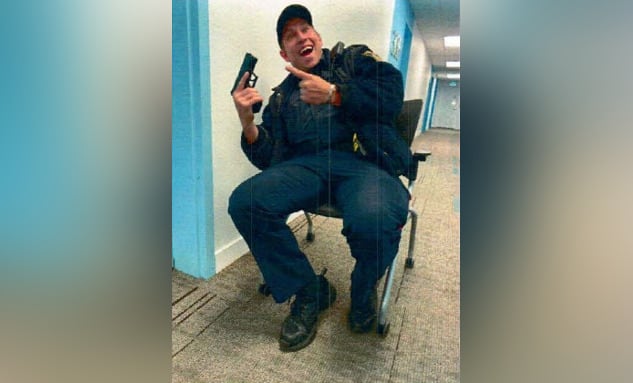 A police officer sitting in a chair in a hallway, holding a gun and smiling.