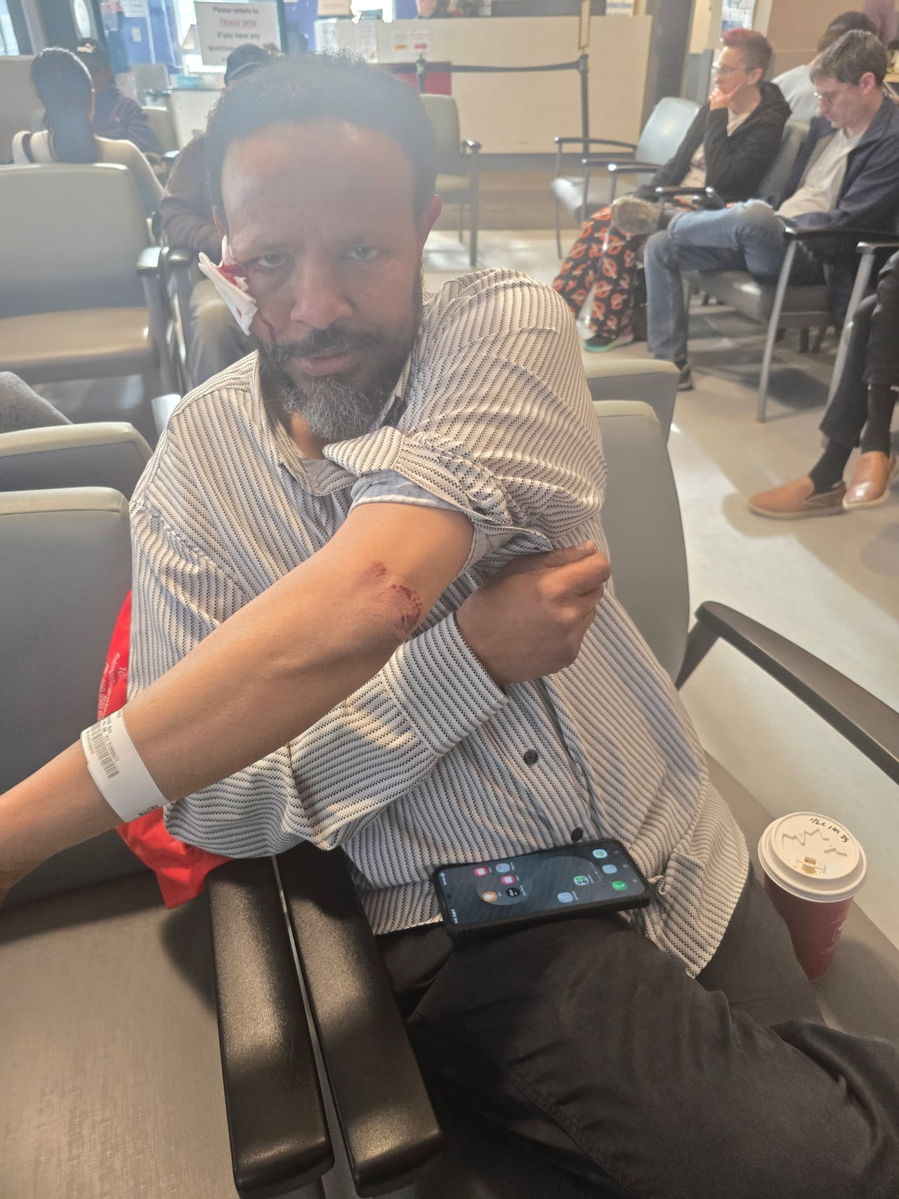 A man sits in a hospital waiting chair with a bandage on his face and a scratch on his elbow