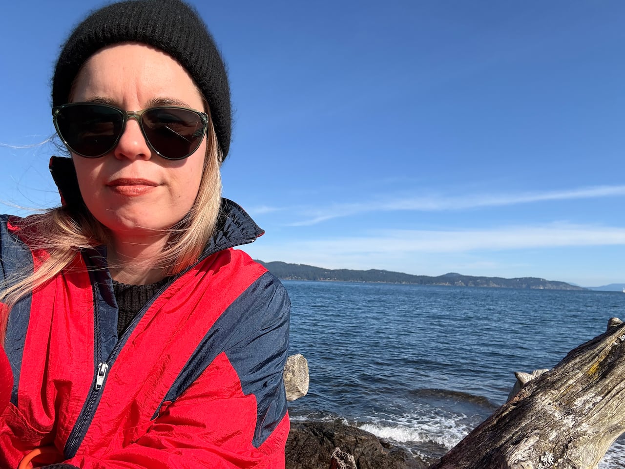 Portrait of a woman in a red and black jacket, toque and sunglasses in front of the ocean.