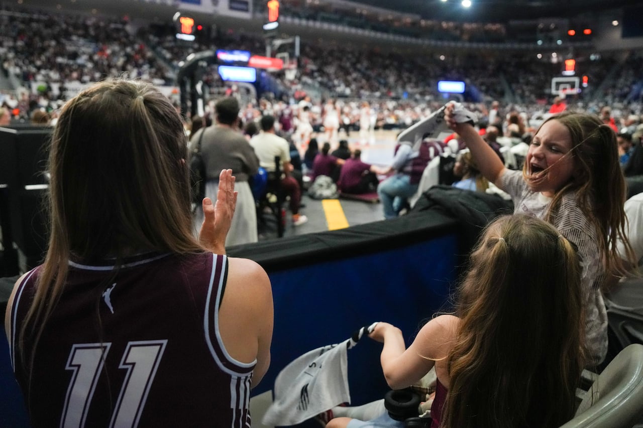 Three girls are shown cheering during a basketball game. The court is in the background while the girls are in the foreground.