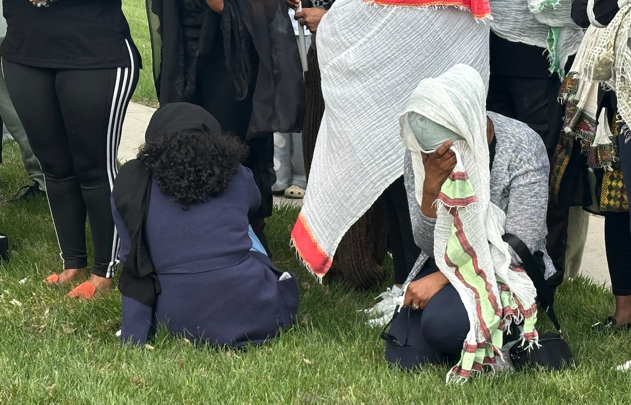 Two woman sit on the grass crying, while other women stand near them holding candles