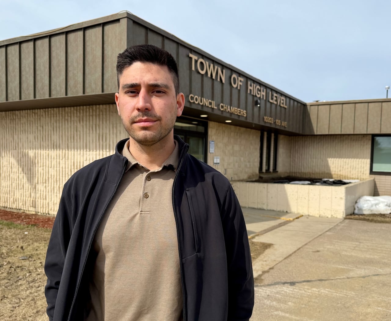 A man in a black jacket and tan shirt stands in front of the town of high level building