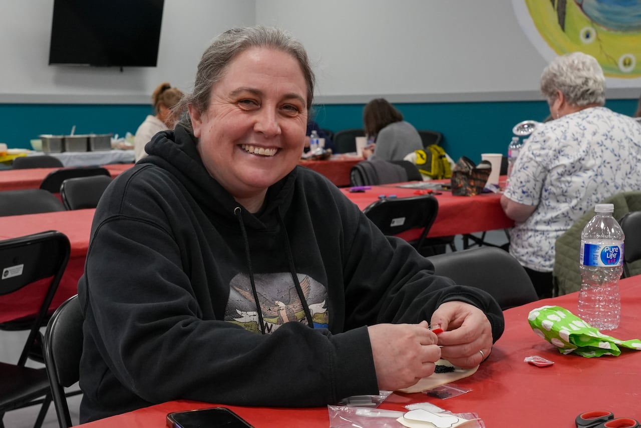 A woman in a black hoodie smiles while sitting at a table covered in arts supplies and a red tablecloth.