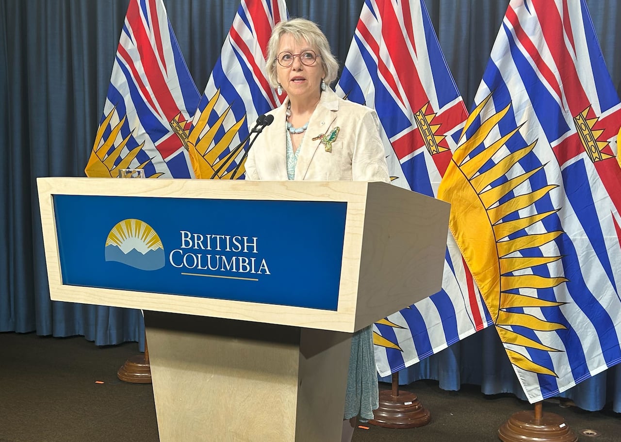 A blonde woman with glasses stands behind a lectern that says "British Columbia" and in front of a row of B.C. flags. She is speaking.