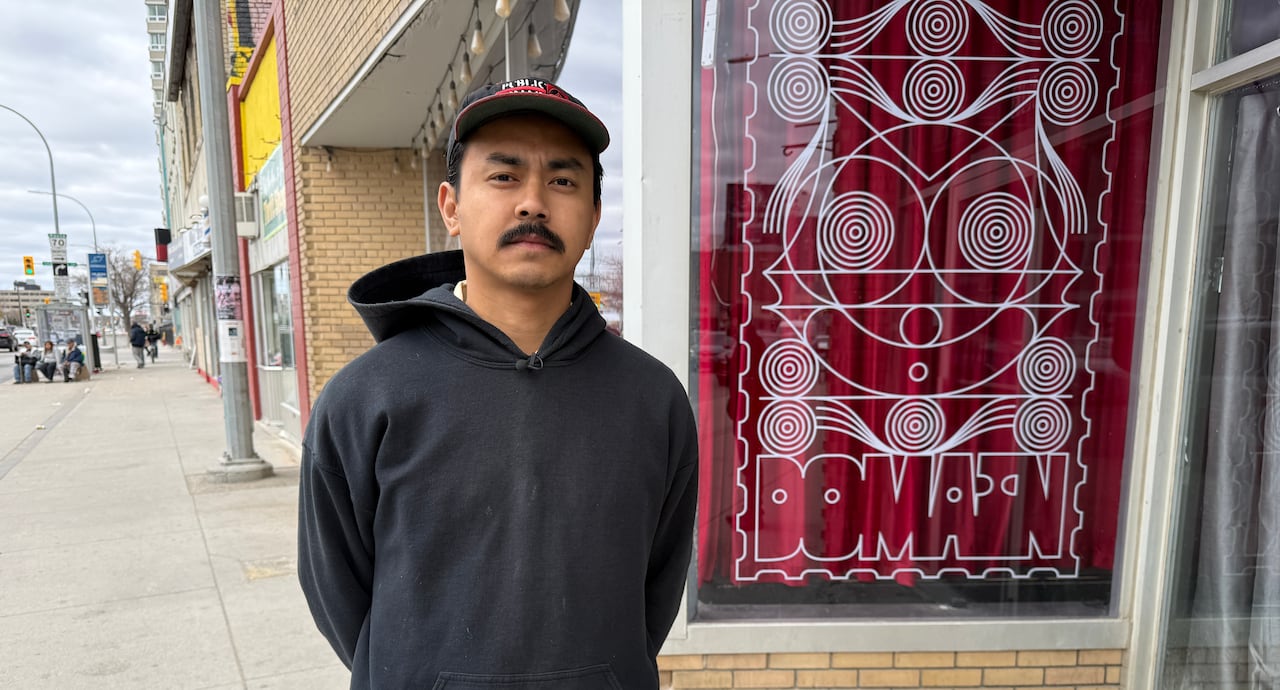Man with dark moustache wears a navy sweatshirt and ballcap, standing outside a storefront along a city street