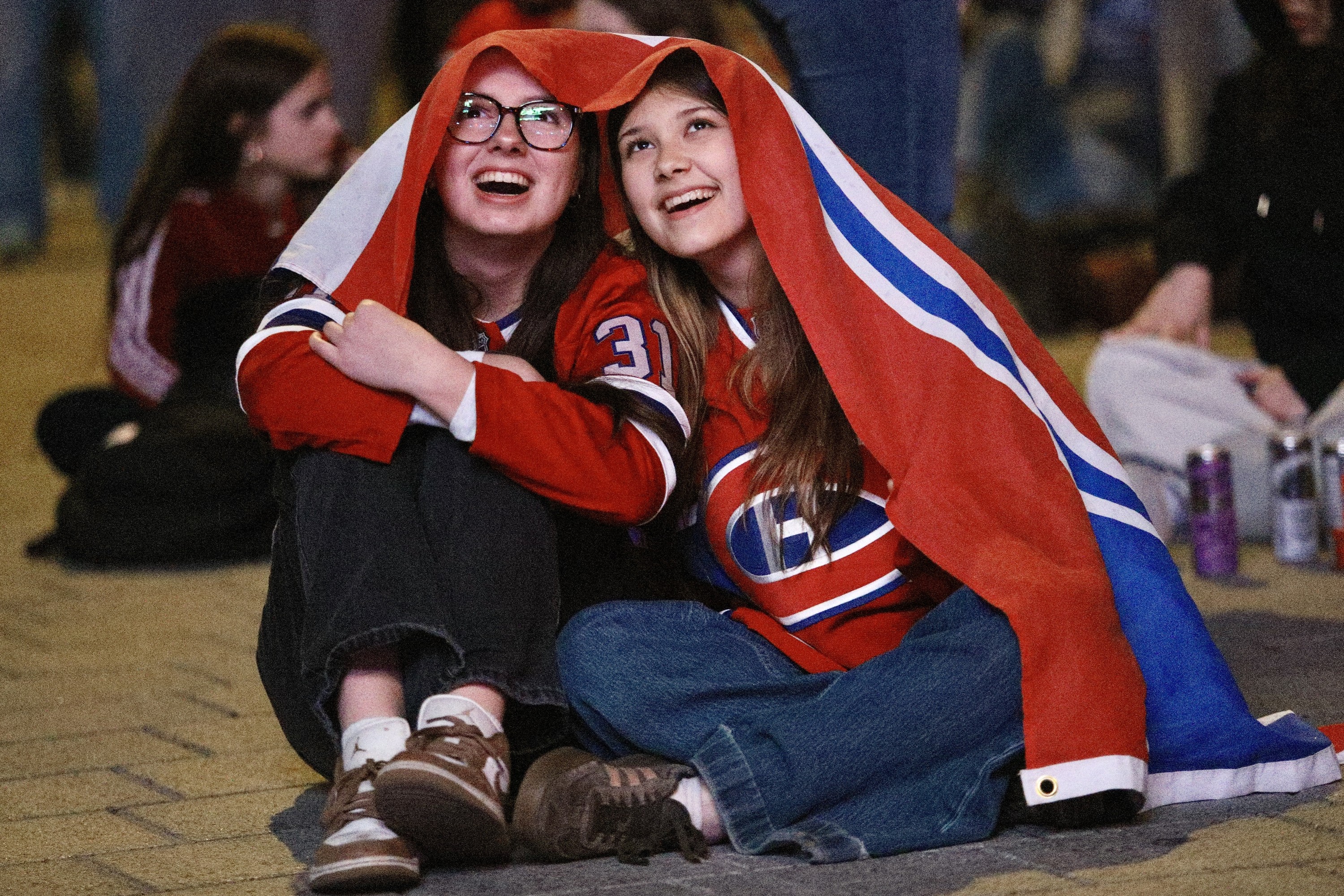 Two young women huddle under a Montreal Canadiens flag outside, smiling as they look into the distance.