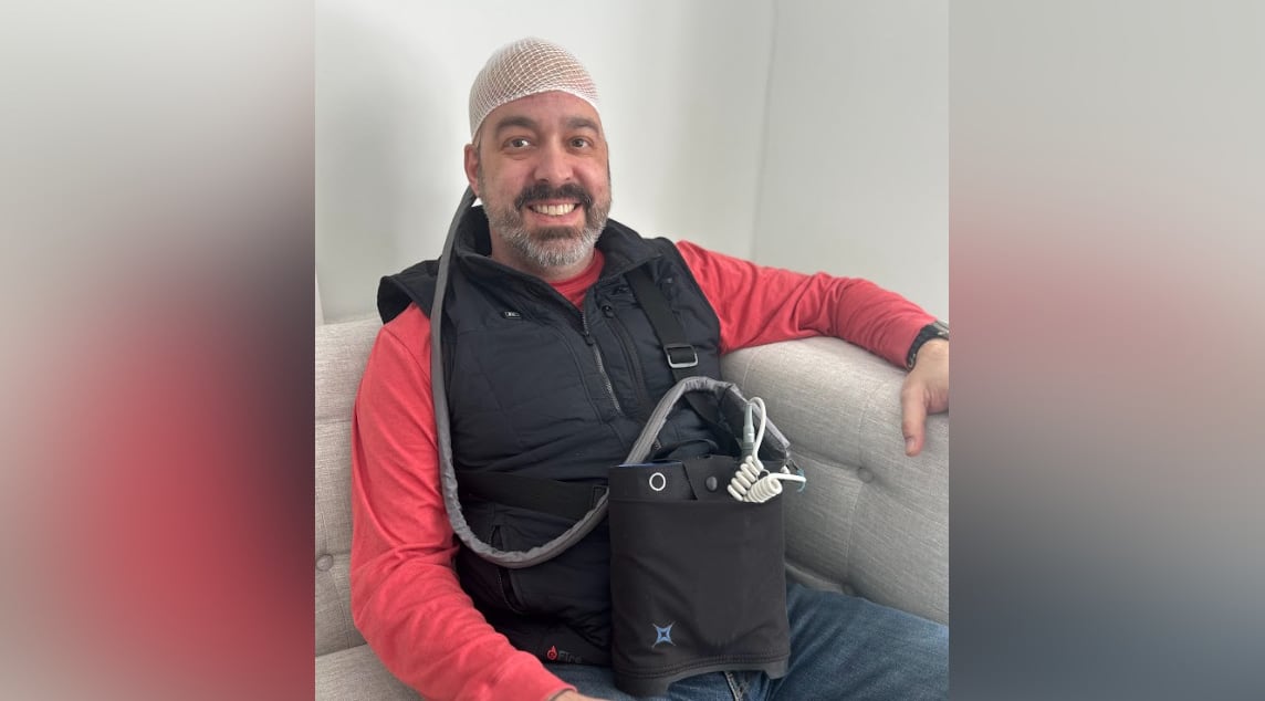 Man smiles and wears netted head cap with tube coming out the back attached to a device in a crossbody bag.