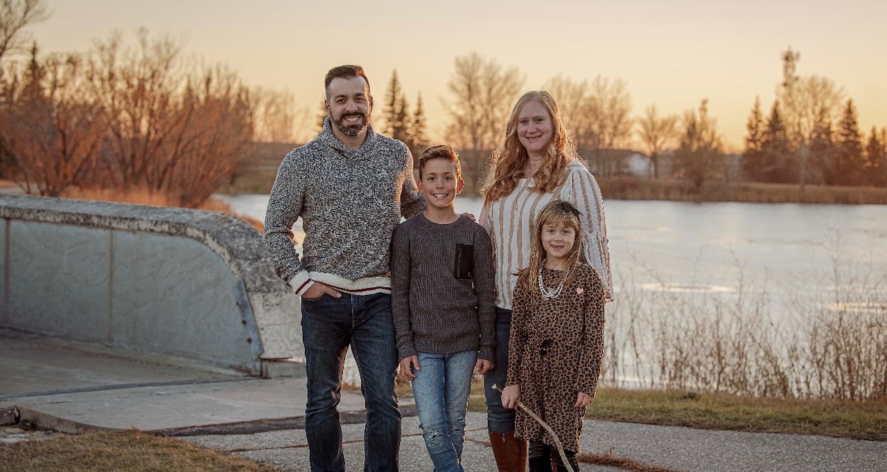Man with dark hair and woman with blonde hair stand with a young boy and a young girl beside a river in a park at sunset
