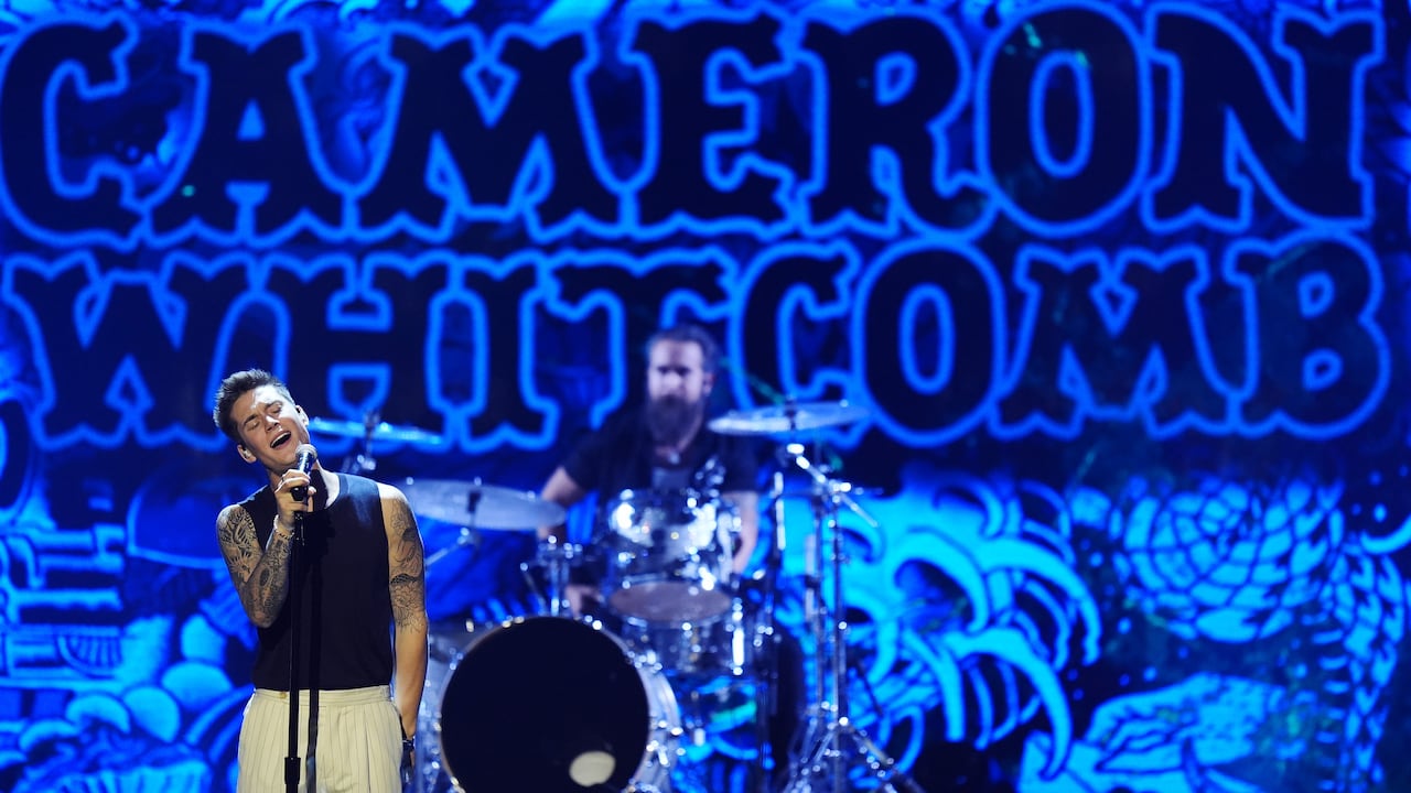 A white male performer stands on stage singing. The name 'Cameron Whitcomb' in blue text is projected behind him.