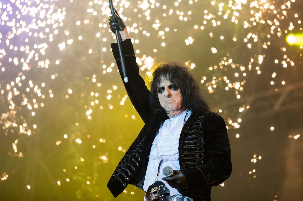 A singer wearing makeup stands in front of sparkles and holds his microphone in the air.