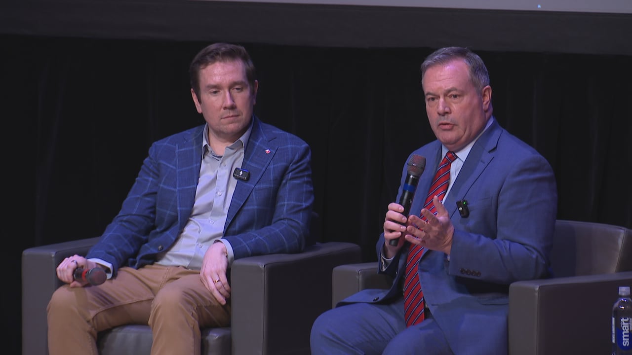 two men in business attire sit in chairs holding microphones as the one on the right speaks