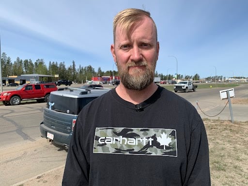 A man with short blond hair and a short beard stands in front of a street. Cars can be seen behind him. He's wearing a black Carhartt shirt. He has a neutral expression on his face.