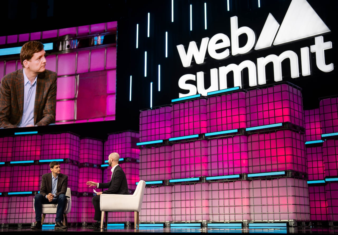 Two men speak at a conference, with one of them projected onto a screen behind him, with the words 'Web Summit' visible.