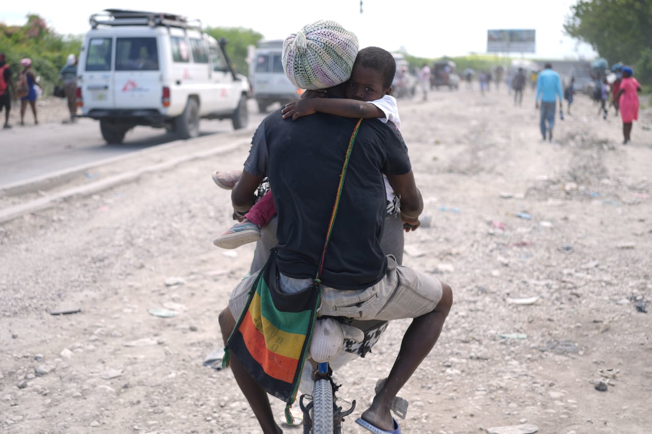 A man riding a bicycle, pictured from behind, with a small child in his lap, arms wrapped around his neck