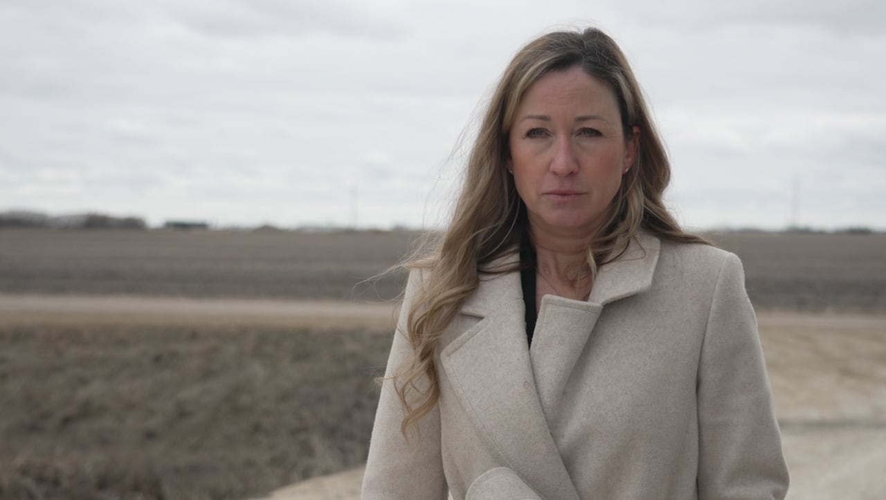 A woman in a tan coat stands in front of an empty farm field in spring