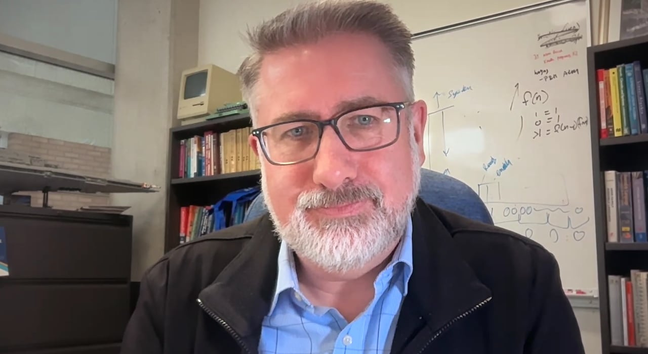 A man with grey hair and glasses sits in an office with bookshelves and a whiteboard