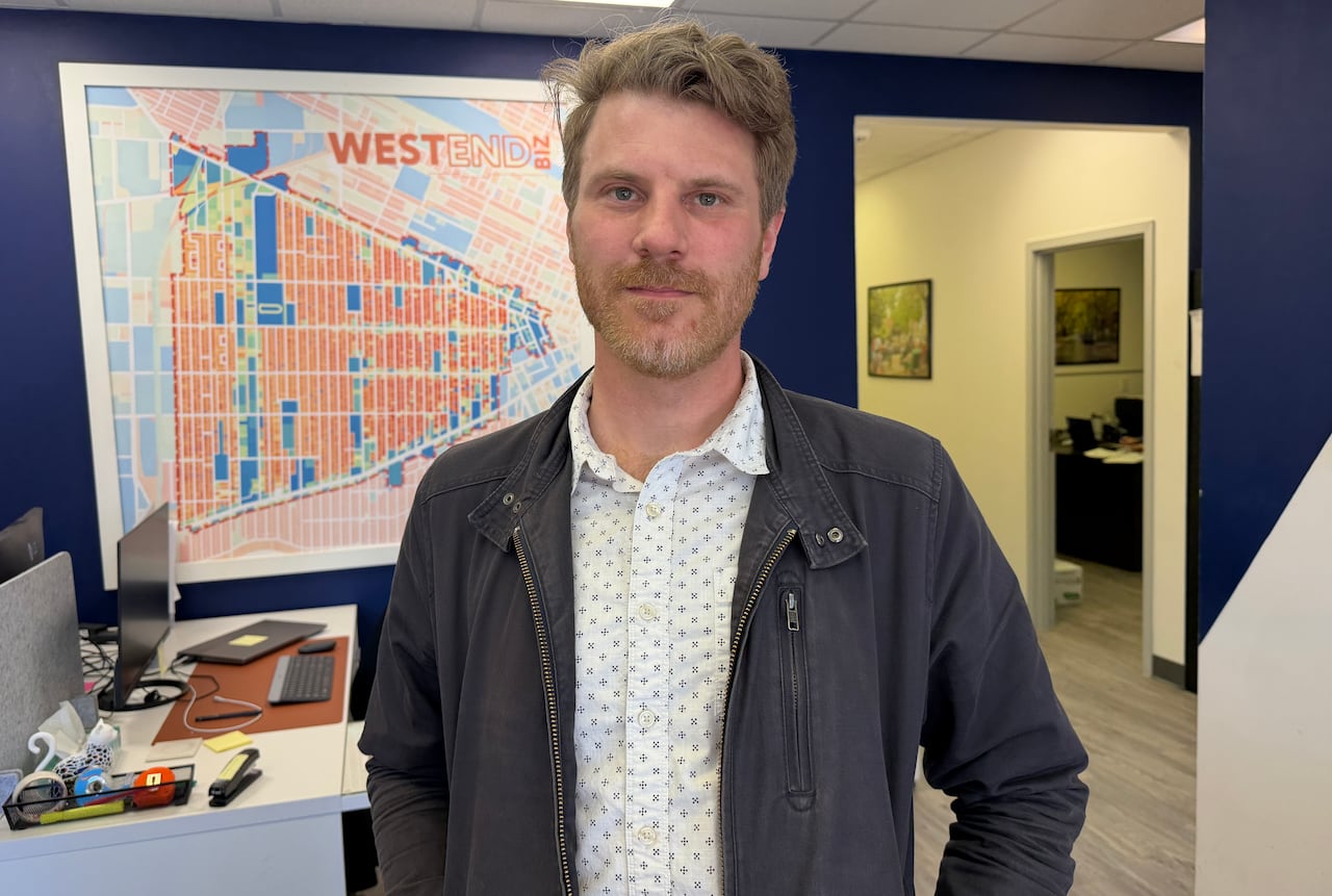 A man in a collared shirt and light jacket stands in an office.