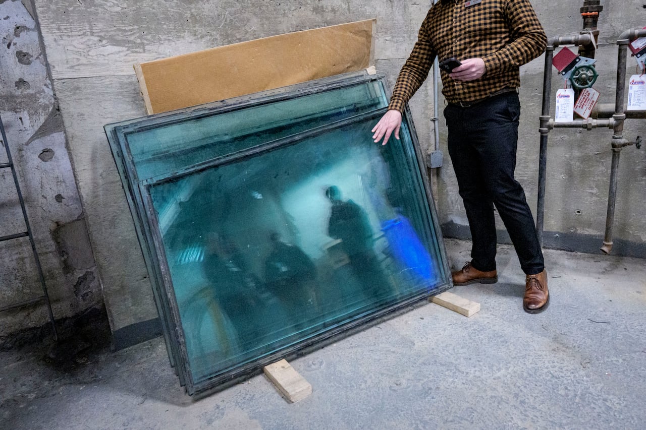 A man pointing at glass panels in a basement chamber, with reflections of several other people in them.