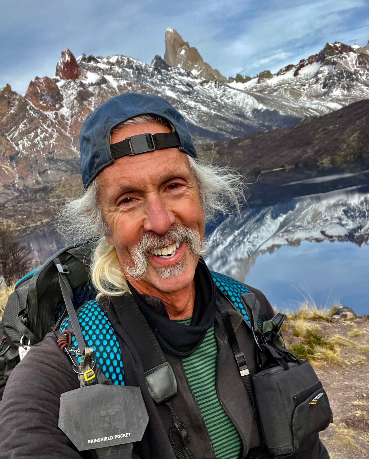 Man with backwards baseball cap poses for selfie in front of a lake mountain side.