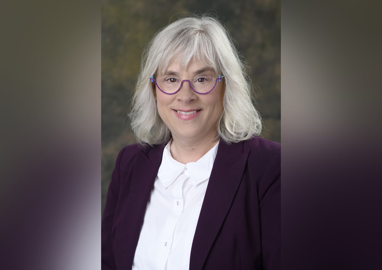 woman with shoulder-length white hair wears purple eyeglasses, a purple blaze and a white collared shirt. 