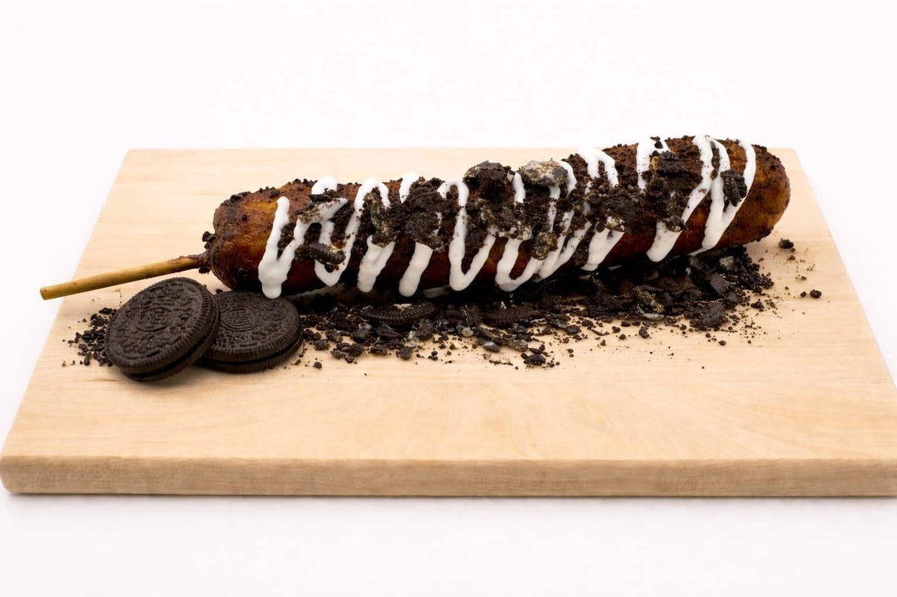 A deep-fried hotdog on a stick, with icing and crushed cookies crumbled on top.