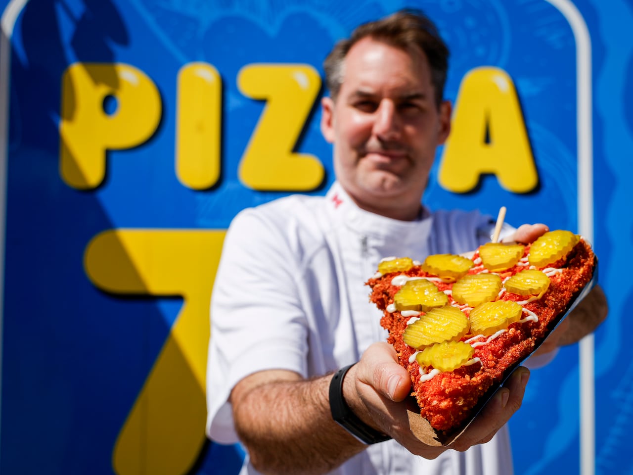 A man holds a slice of deep-fried pizza with pickles on it up for the camera.