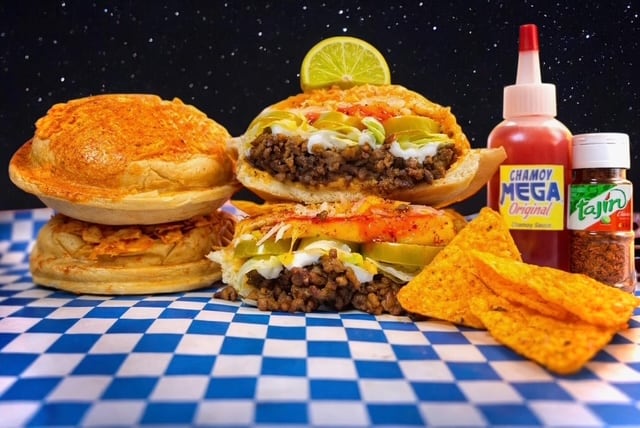 A stuffed burger is cut in half with a bottle of sauce and potato chips sitting next to it.