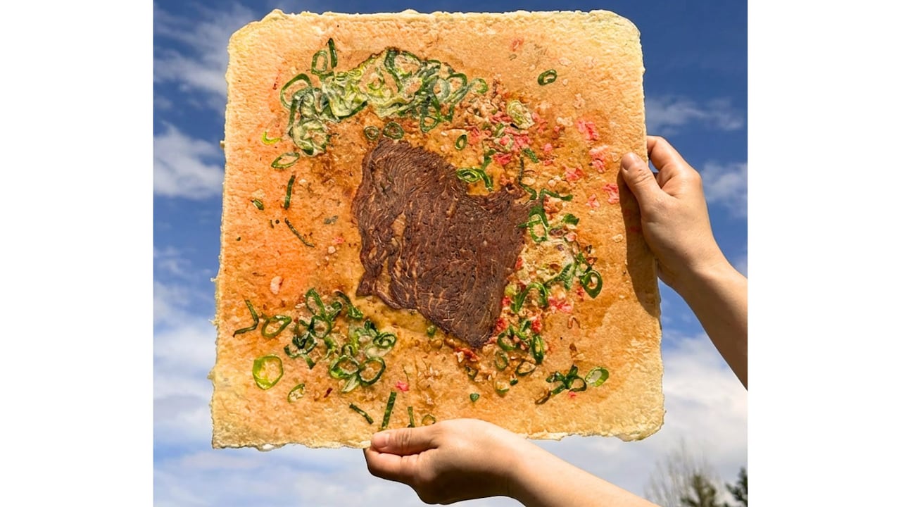 A person holds up a giant grilled rice cracker with a slice of beef in the centre.