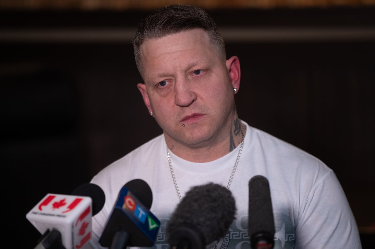 A man wearing a white T-shirt sits in front of a number of microphones.