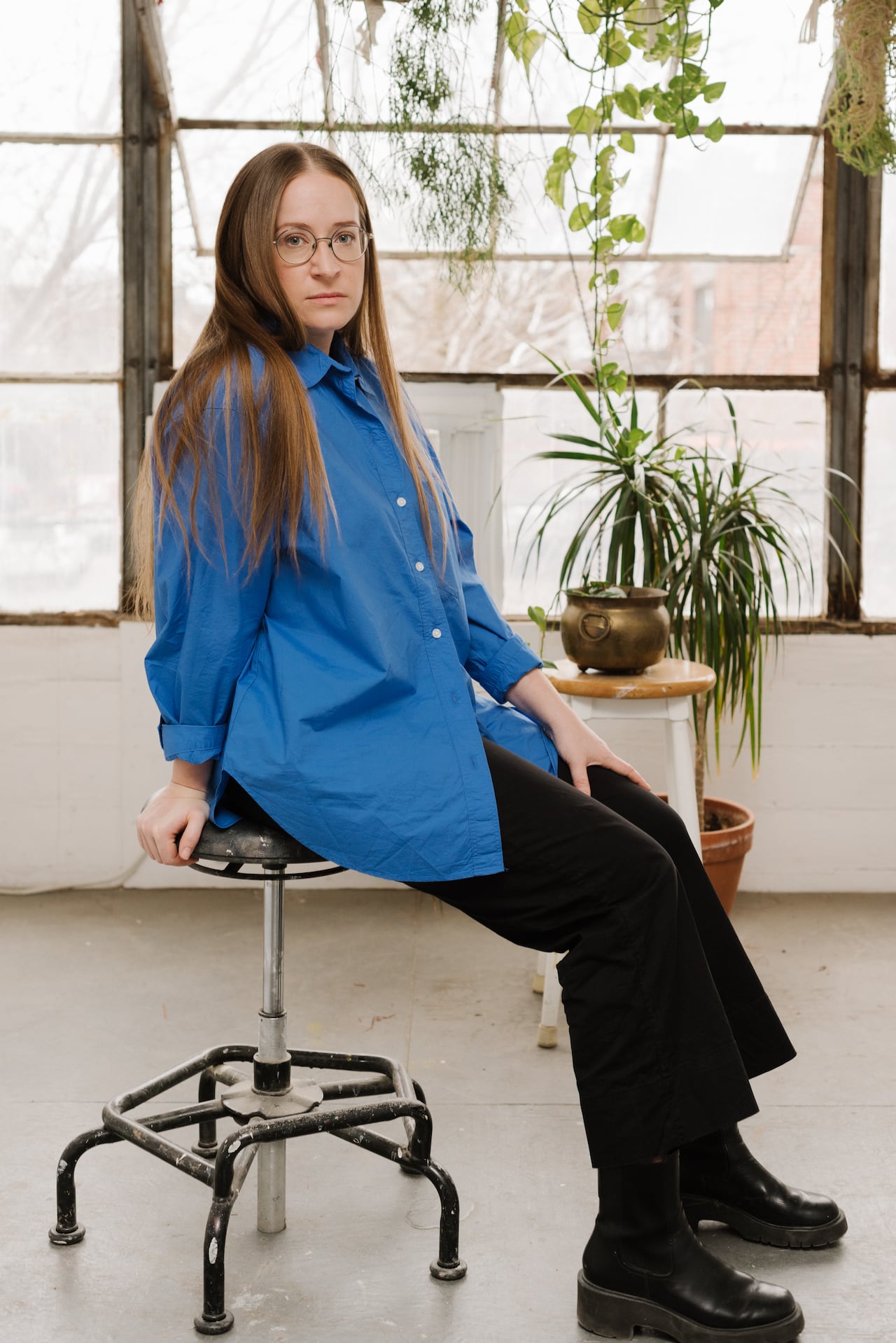 A woman with long, reddish blond hair and glasses wearing a blue button up and black pants sits on a stool in front of a large glass window with houseplants.