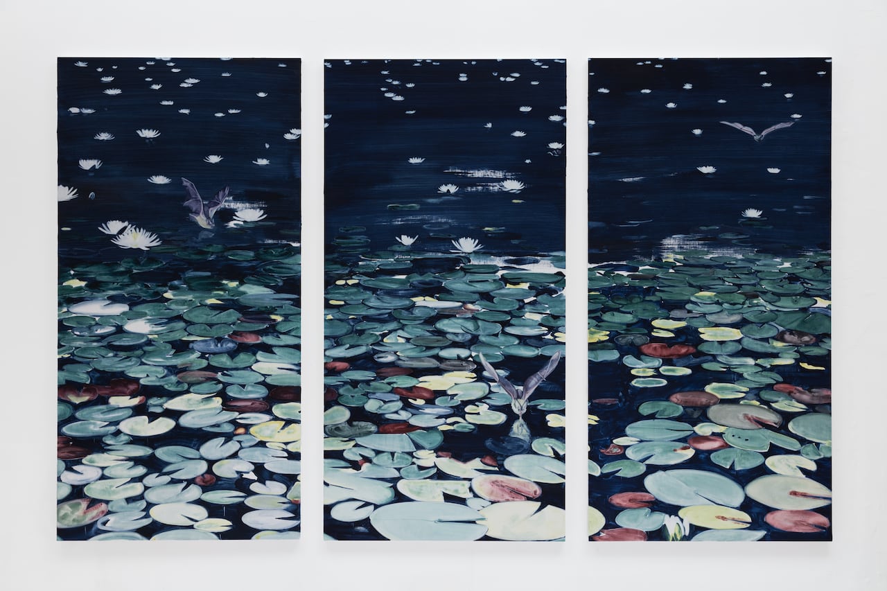 Three panels hang beside each other on a white wall. The paintings depict a nighttime scene, showing a pond covered in lily pads and flowers. Three bats swoop around the scene.
