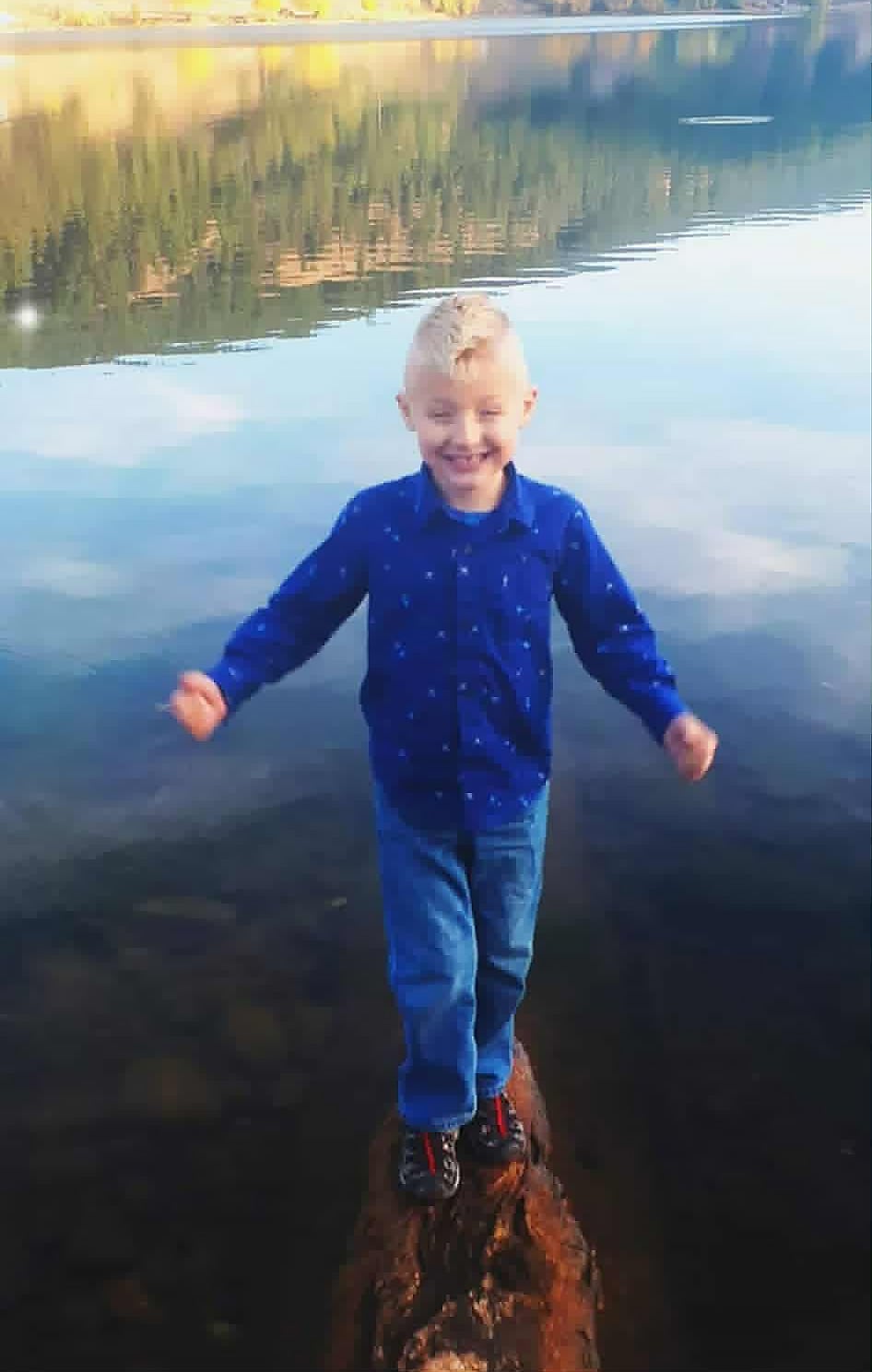 A boy with blond hair stands in front of a body of water.