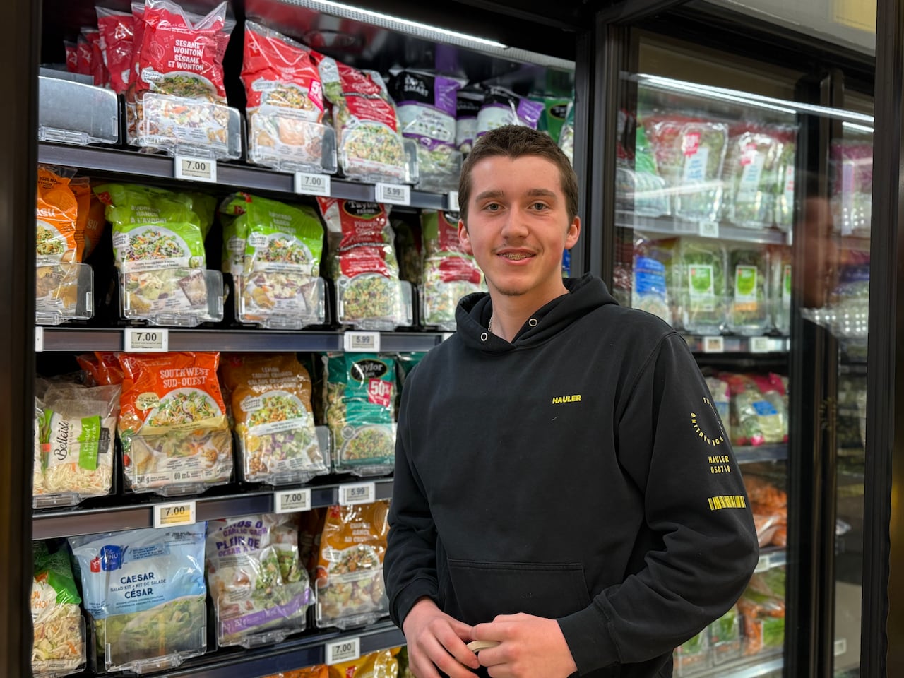 A young man in a black hoodie stands smiling in front of a refrigerated grocery store shelf stocked with various packaged salad kits
