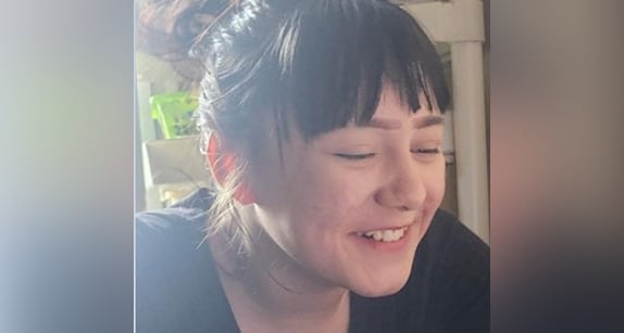 Young girl with dark hair and bangs in a ponytail is shown from the side, wearing a black shirt and smiling