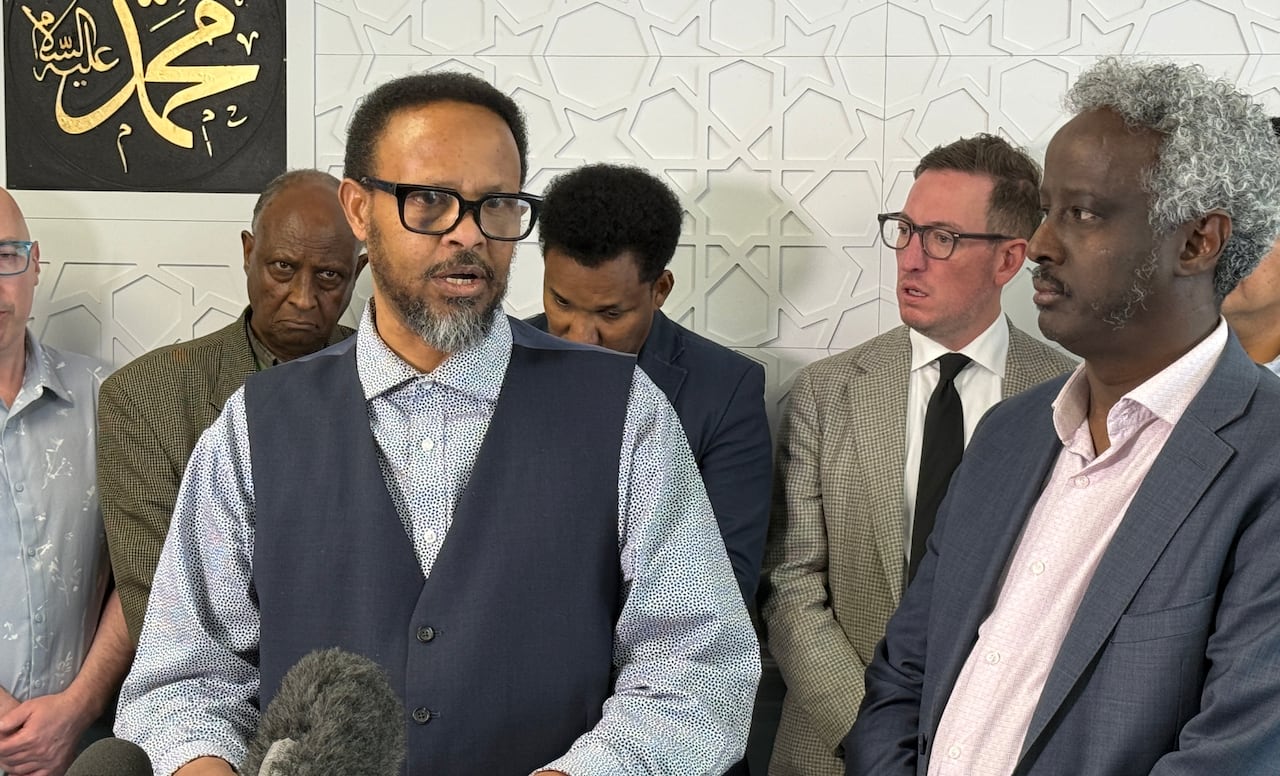 Two Black men are standing at a podium. One of them, wearing thick-rimmed glasses and a vest over a dress shirt, is speaking into a microphone. They stand in front of a lineup of white and Black men.