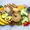 Overhead view of a large group of food with high content of dietary fiber arranged side by side. The composition includes blueberries, oranges, avocado, wholegrain bread, potatoes, oats, corn, beans, greens, broccoli, mixed nuts, and bananas, among others.