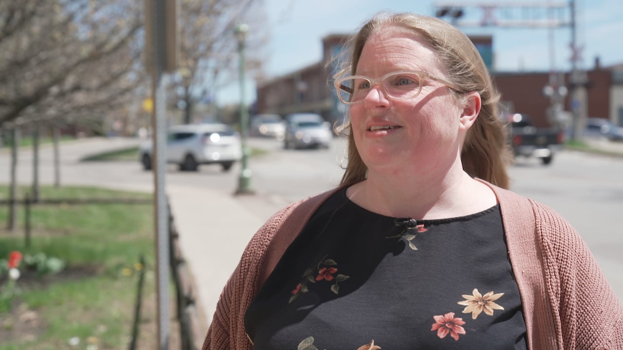 A woman with long brown hair stands in front of Main Street Sussex. She is wearing clear glasses, a floral shirt, and a pink cardigan.