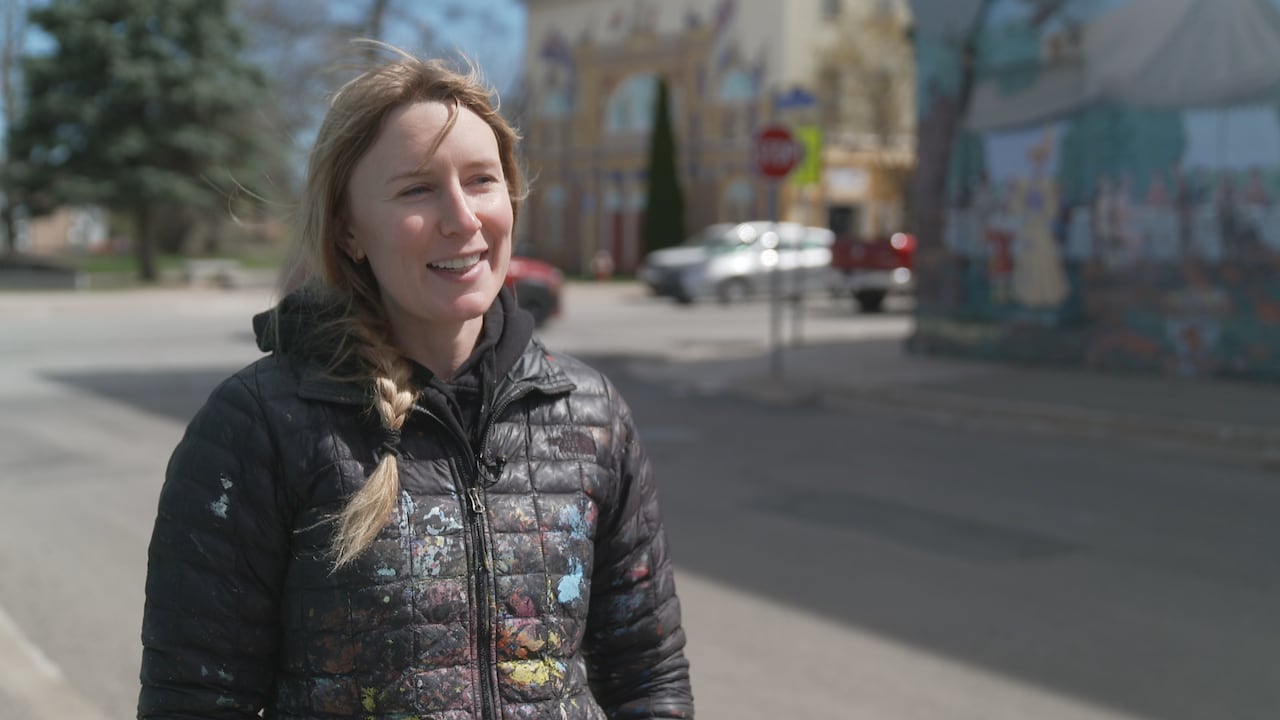 A blonde woman with a braid, wearing a black jacket splattered in paint. She is taking a break from painting a mural.
