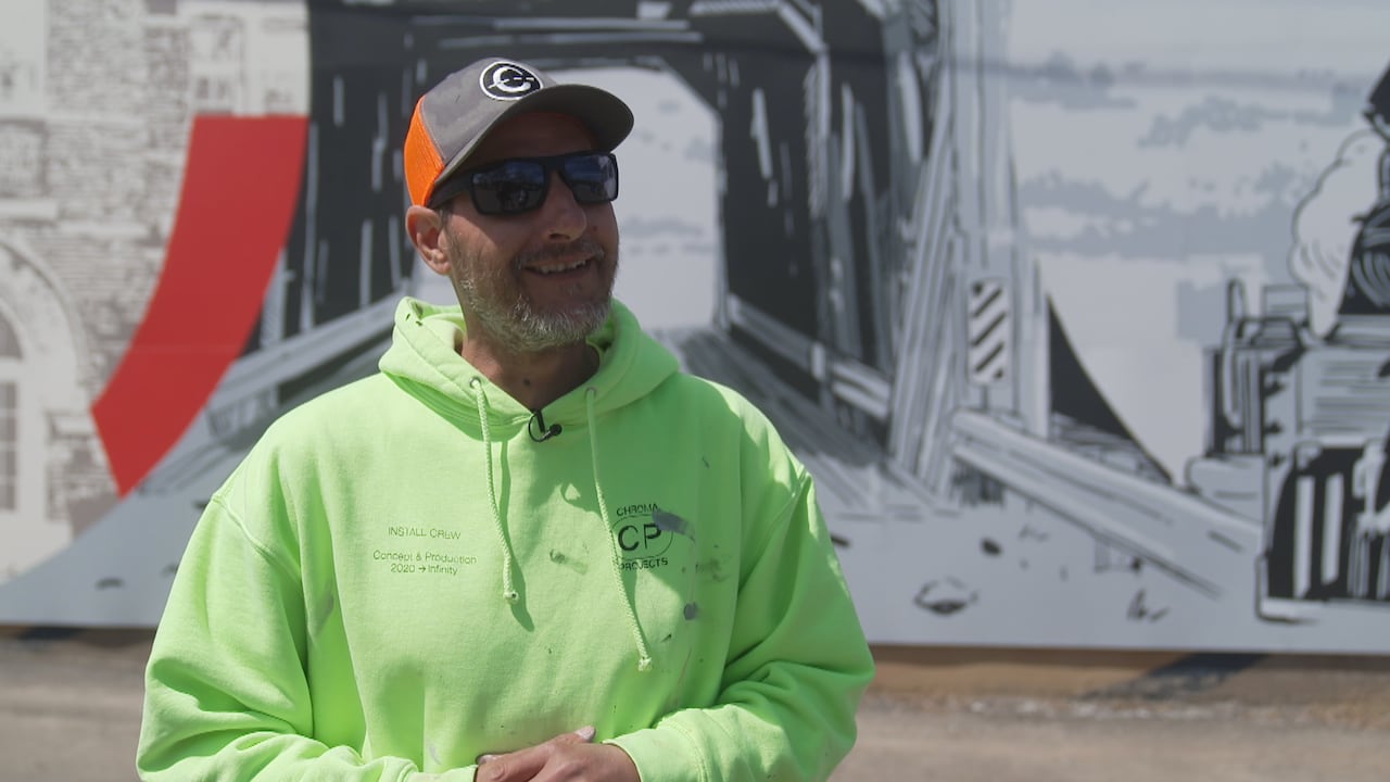 A man wearing sunglasses, a baseball cap, and a neon green hoodie speckled in paint, stands in front of a mural he has just finished.