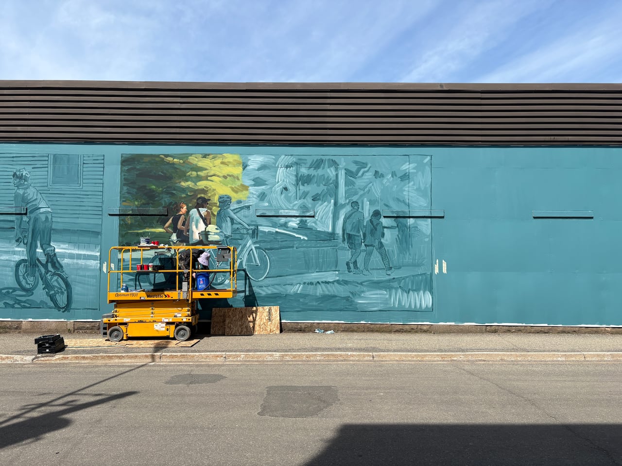 A painter crouches down on a yellow cherry picker in front of a massive blue outline of a mural, depicting different scenes of people on bikes.