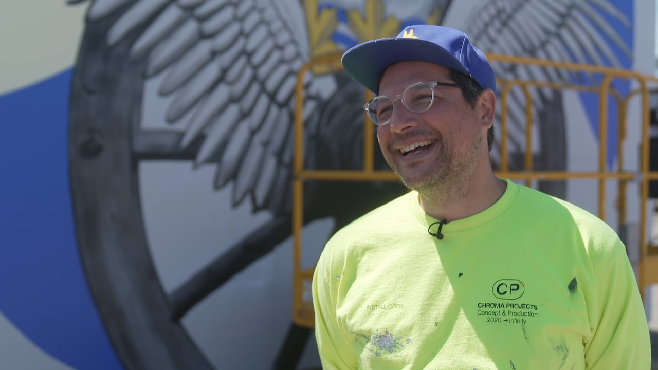 A man wearing clear glasses and a blue baseball hat and a neon green shirt splattered in paint stands in front of a finished mural.