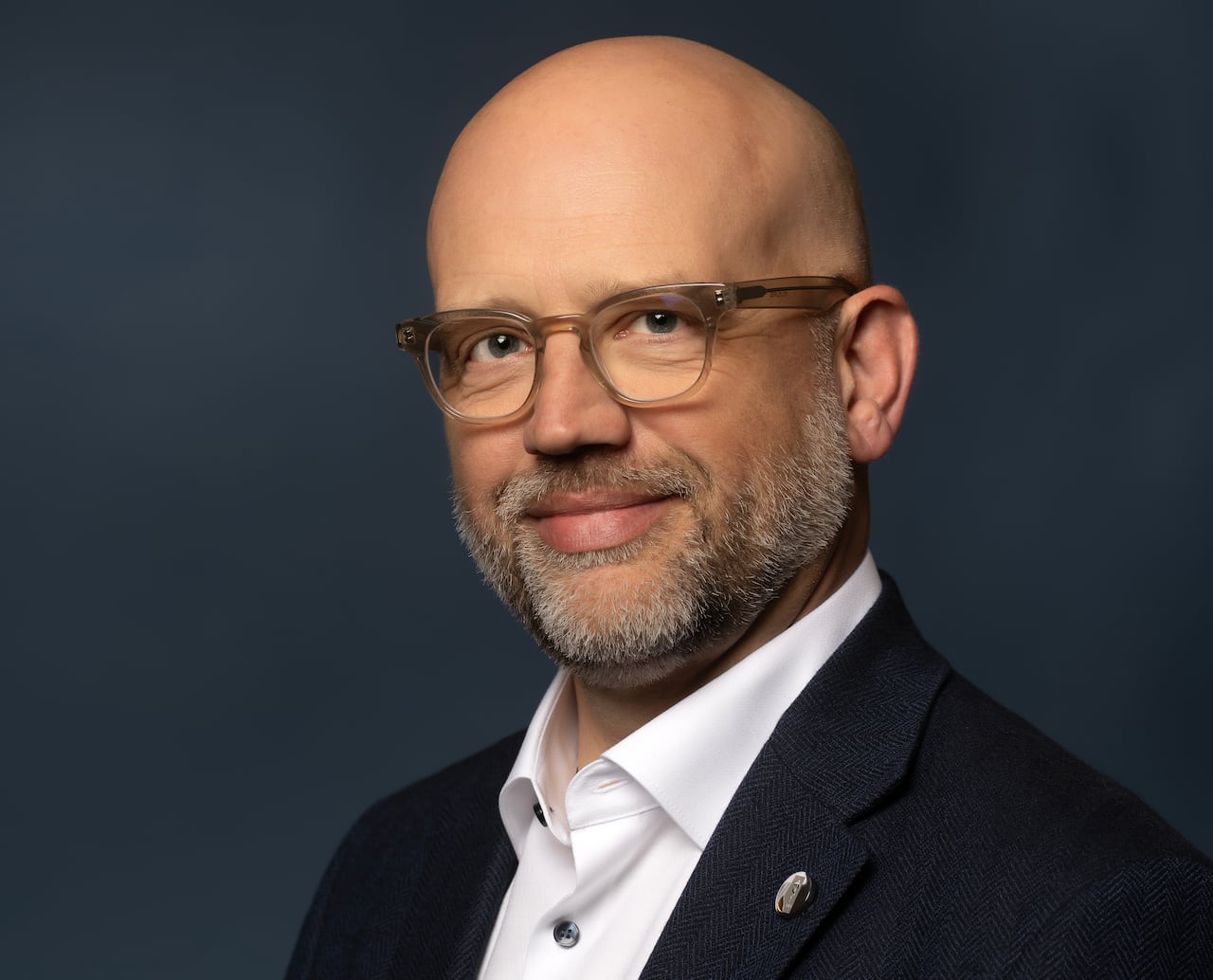 A man with a shaved head, glasses and beard is wearing a black suit and white shirt and smiling.