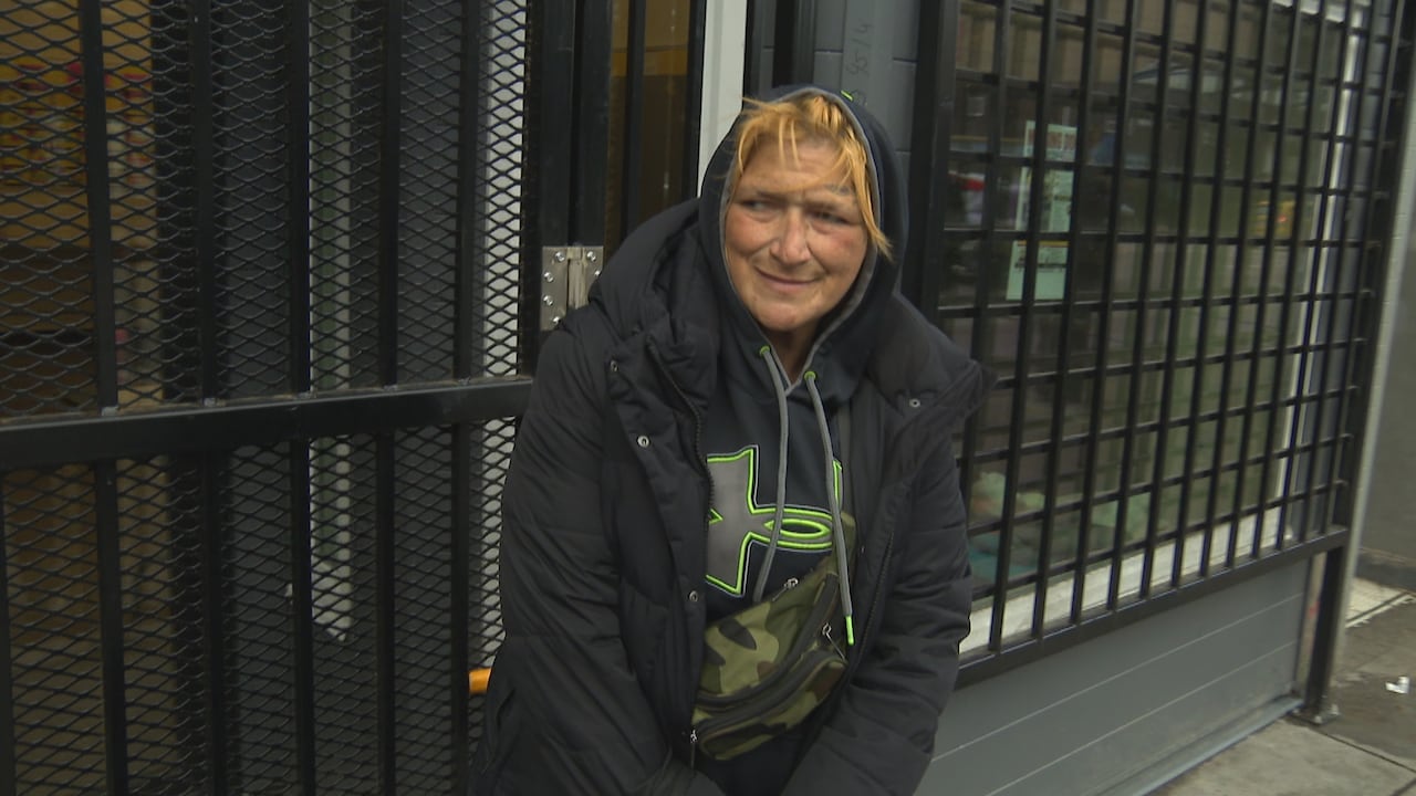 A woman wearing a black jacket and hoodie sits outside a building with metal bars on the windows.