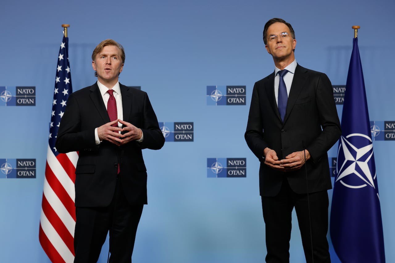 Elbridge Colby and Mark Rutte stand in front of a blue backdrop with the NATO emblem, flanked by U.S. and NATO flags.