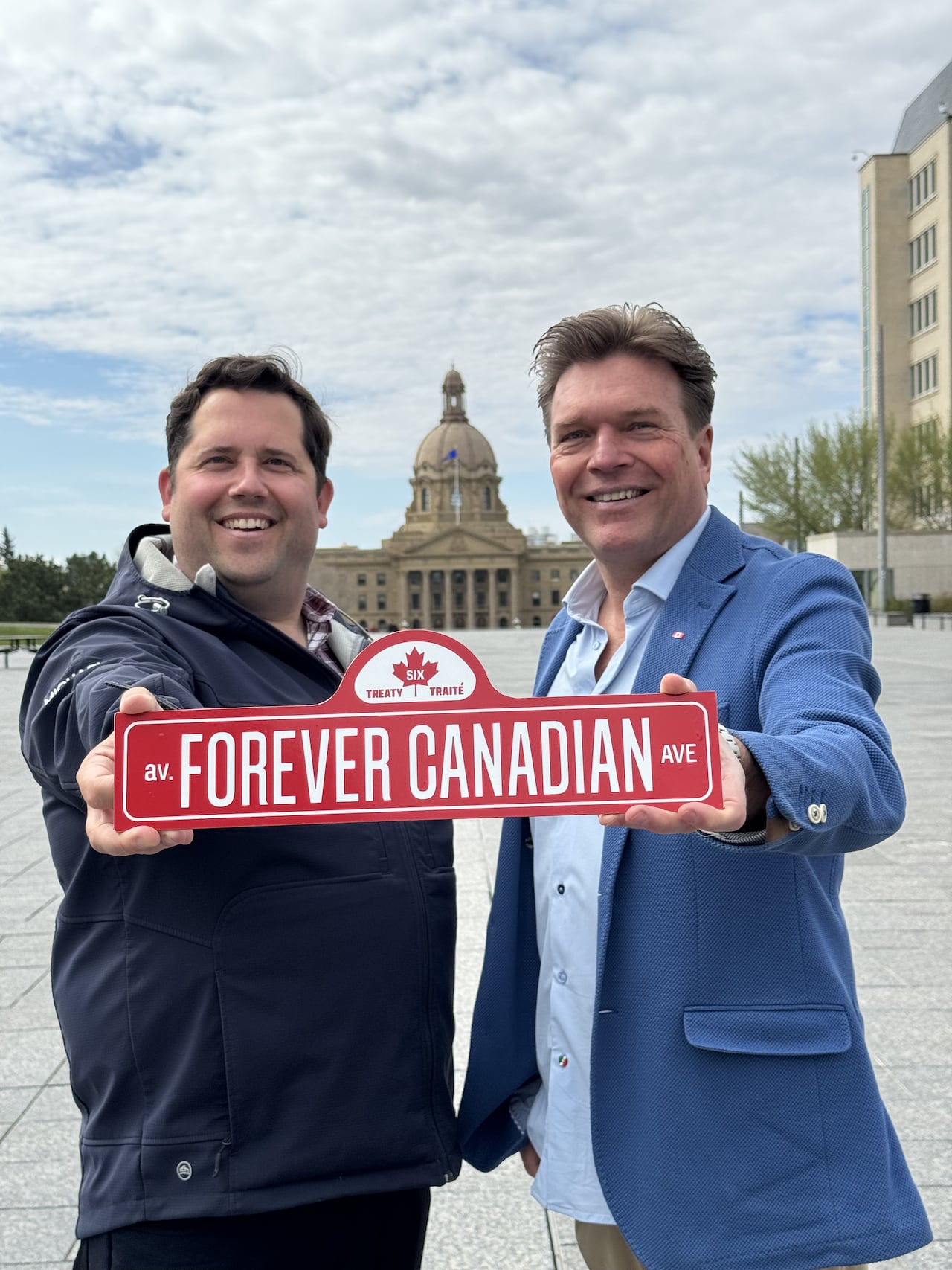 Two men hold a red-and-white sign with a maple leaf at the top, reading, "av. Forever Canadian Ave." The Alberta legislature building is visible in the background.