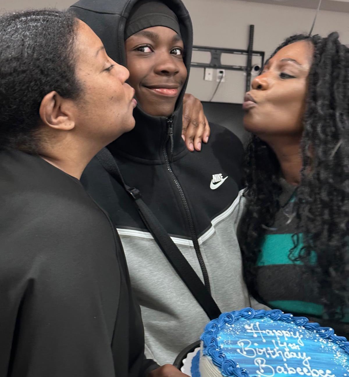 A Black man in a grey and black hoodie stands holding a white cake with blue icing. Two Black women stand on each side of him as they both give him a kiss on the cheek. 