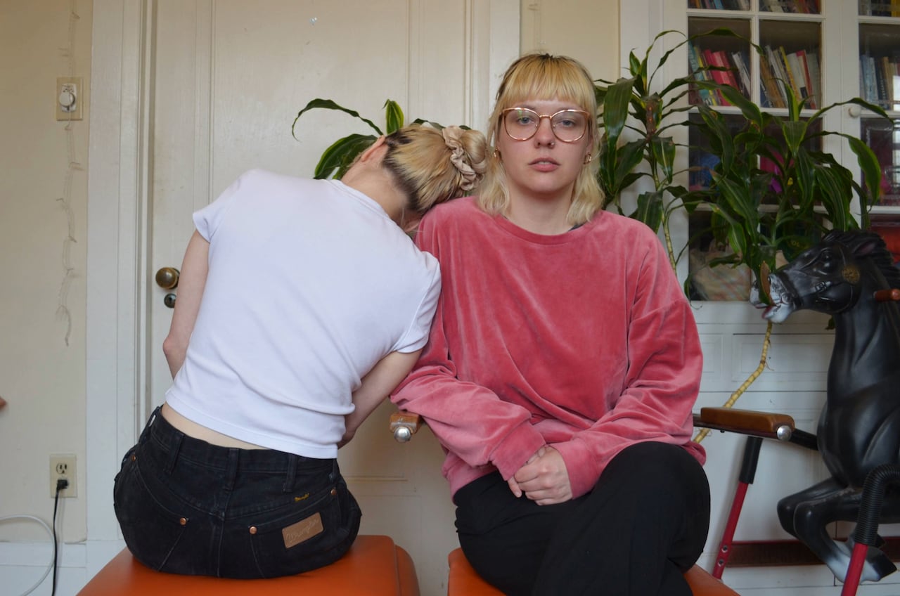 A woman in a pink sweater faces the camera. Another woman leans her head on the shoulder facing away from the camera.