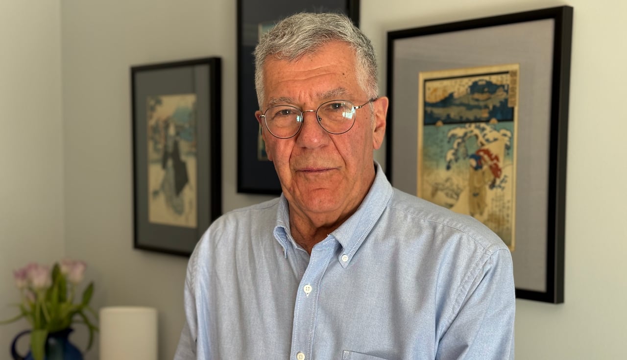 Sandford Borins, wearing a button up shirt and glasses, stands in front of a wall with art work hanging.