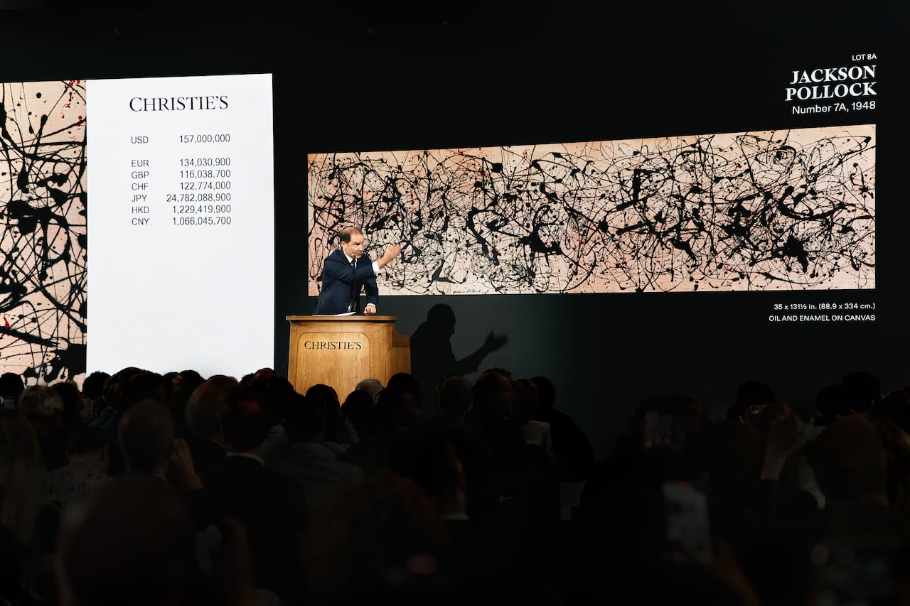 A man at a spotlit podium gestures to the right, an abstract artwork projected behind him, as an audience sits in darkness below him.