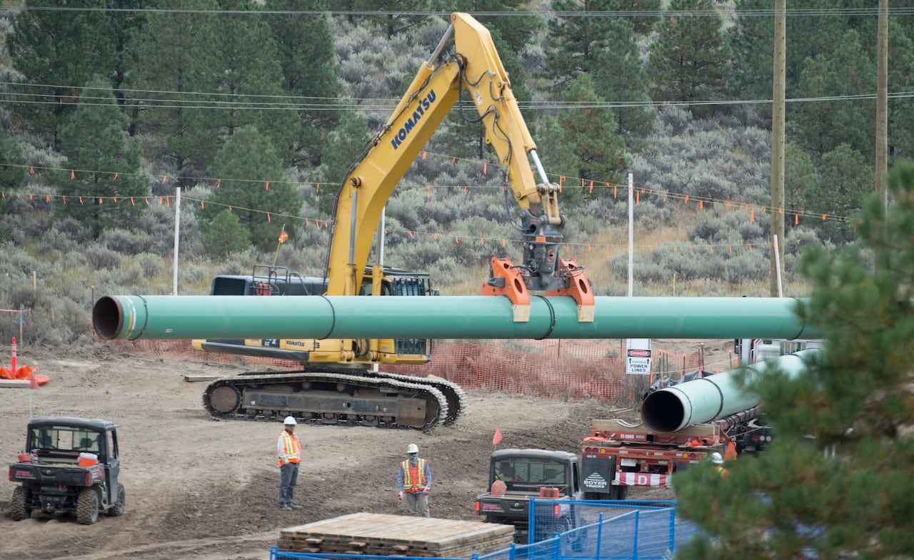 A yellow construction vehicle lowers a green pipeline into a ditch near two other construction workers.